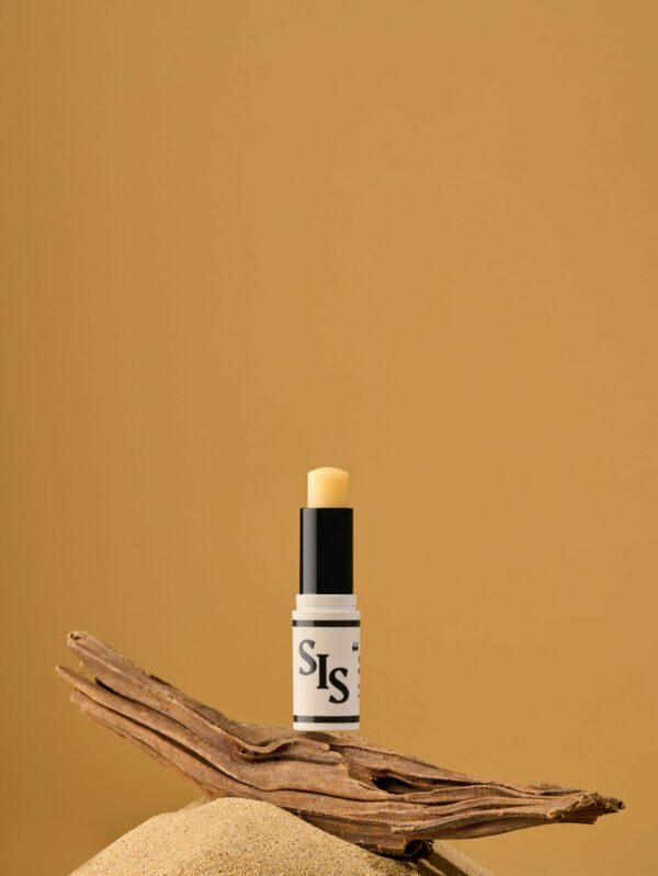 SIS, Romance en Calabre le parfum solide n°2. Stick crème avec logo et ligne noire. La formule est de couleur jaune pour rappeler le soleil. Le stick est ouvert sur une branche de bois sur une petite montagne de sable. Le fond est jaune. Pour rappeler un été au soleil!