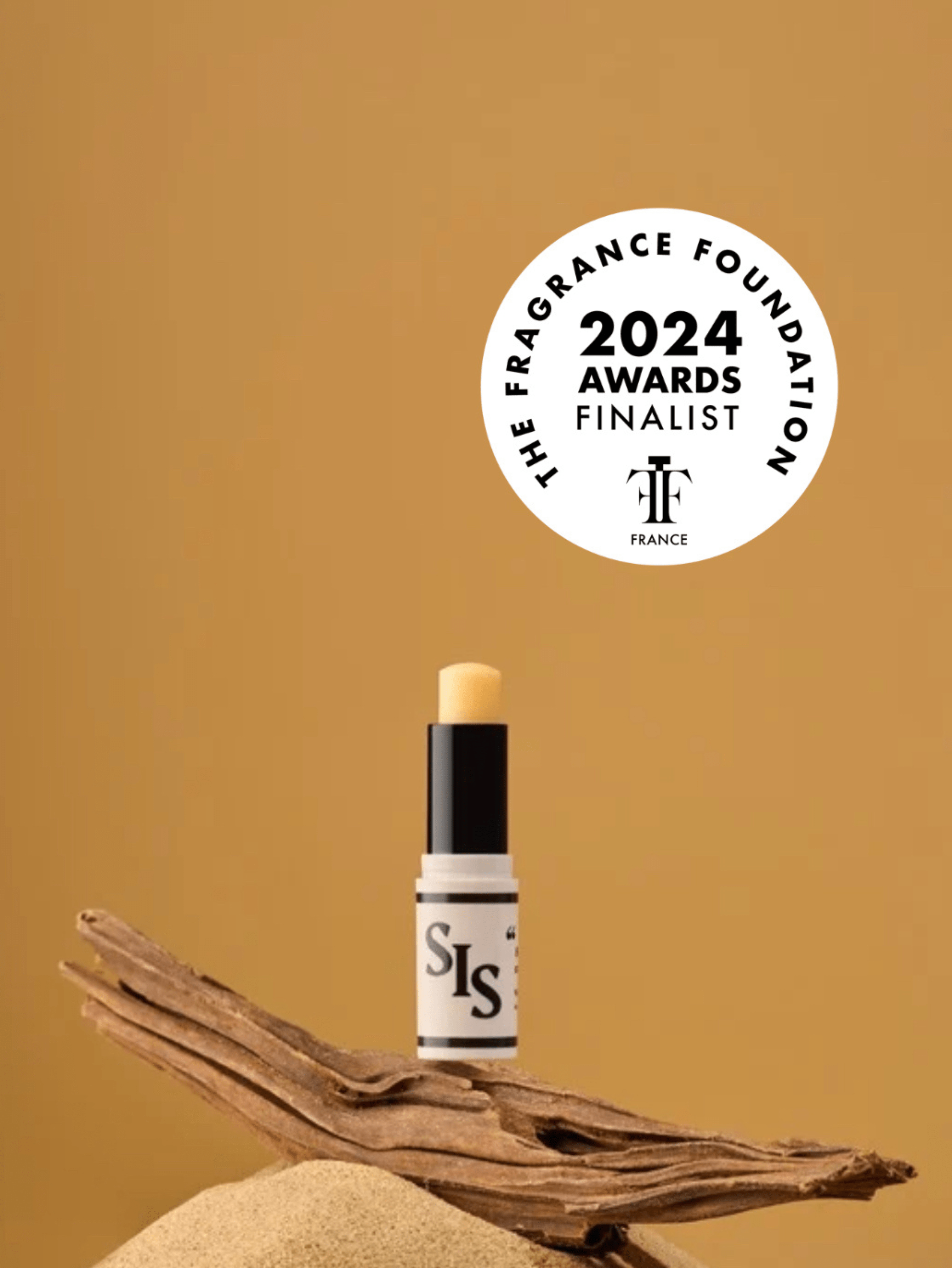 SIS, Romance en Calabre le parfum solide n°2. Stick crème avec logo et ligne noire. La formule est de couleur jaune pour rappeler le soleil. Le stick est ouvert sur une branche de bois sur une petite montagne de sable. Le fond est jaune. Pour rappeler un été au soleil!